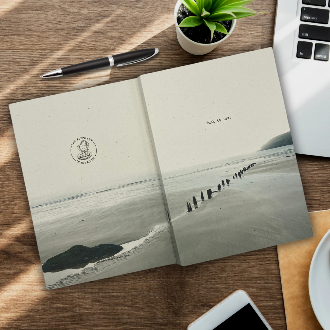 fuck it list notebook with a beach scene spanning from front cover to back. Open on a desk