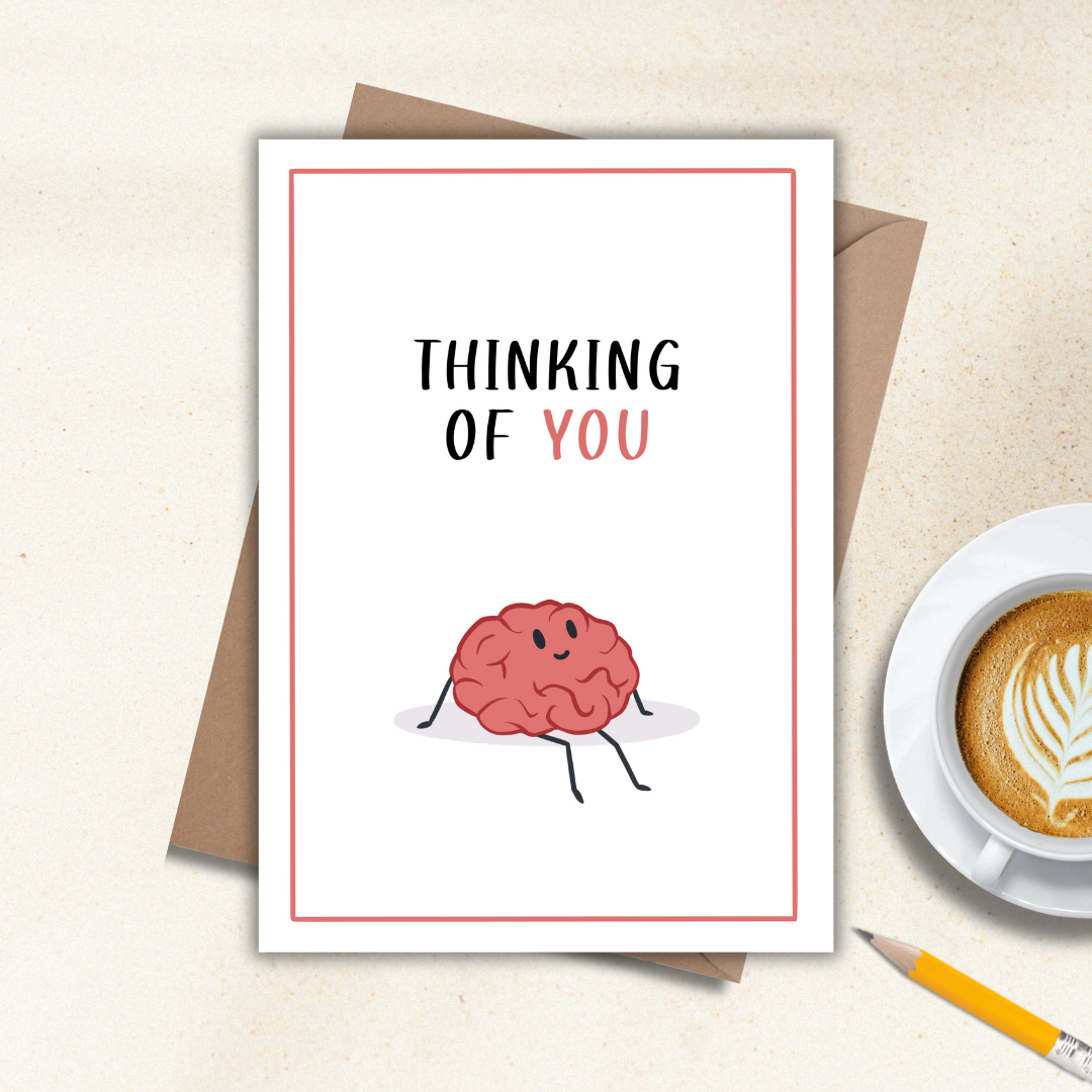 Thinking of you card with a cartoon brain and 'Thinking of You' text, placed on a surface with a cup of coffee and pencil.