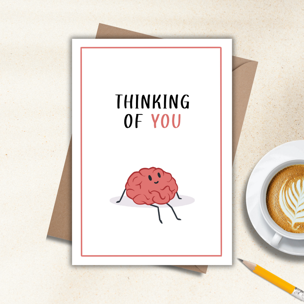 Thinking of you card with a cartoon brain and 'Thinking of You' text, placed on a surface with a cup of coffee and pencil.